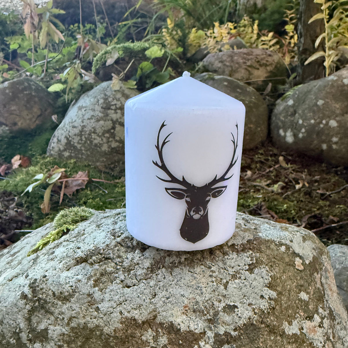 A white pillar candle with a black deer head graphic sits on a large mossy rock in an outdoor garden setting.